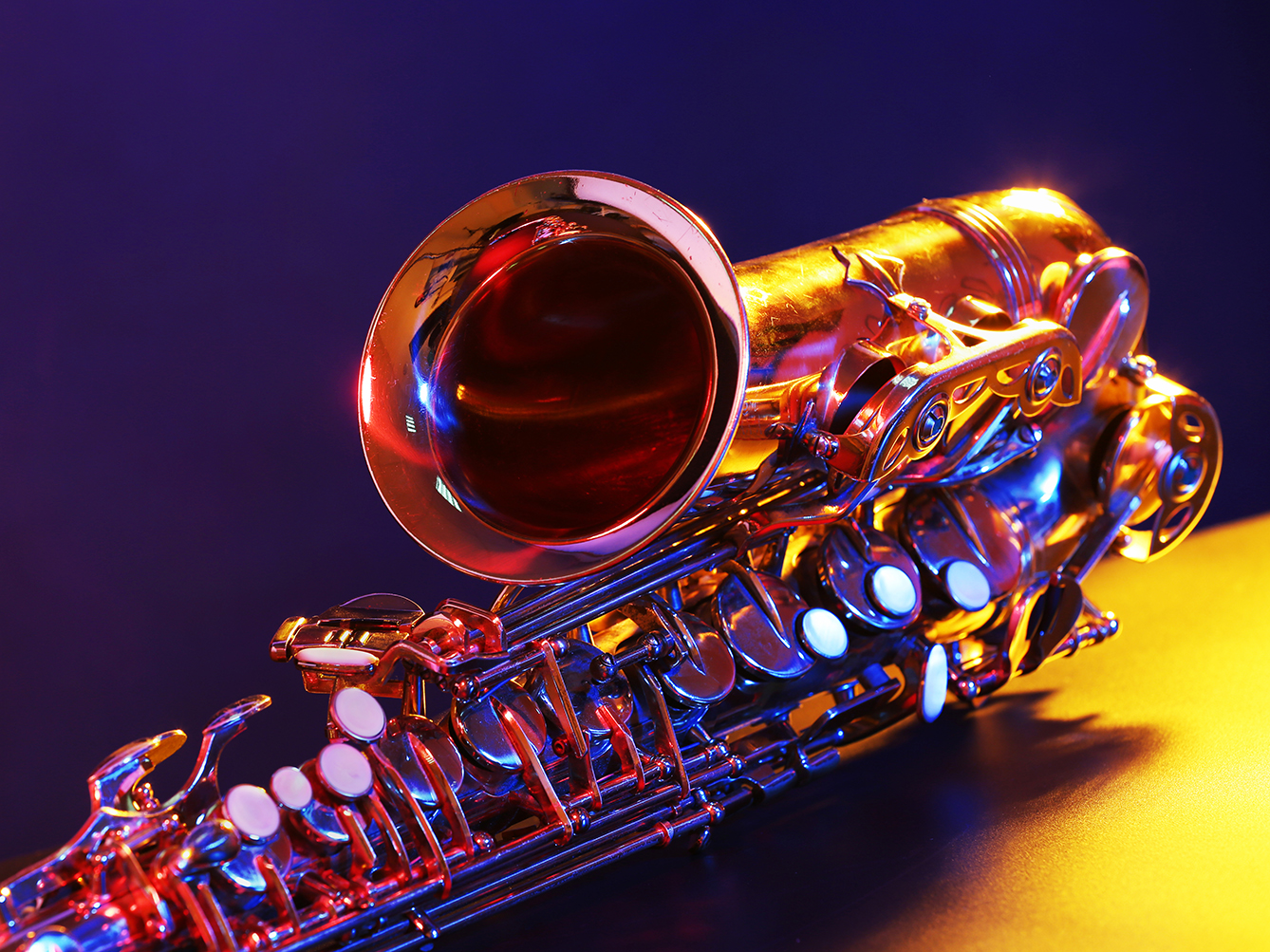 Photo of a saxaphone on it's side with colourful lights reflected on the shiny surface. The background is Alternating solid yellow and purple.